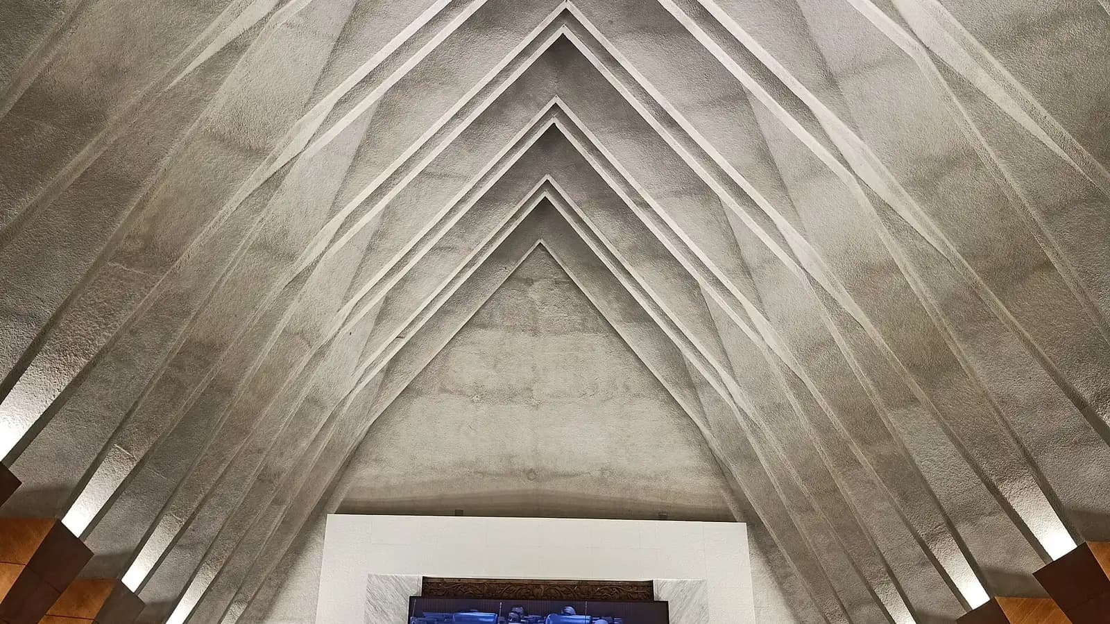 The ribbed, triangular ceiling of the Dewan Rakyat chamber, with a screen showing the sitting at the bottom of the frame