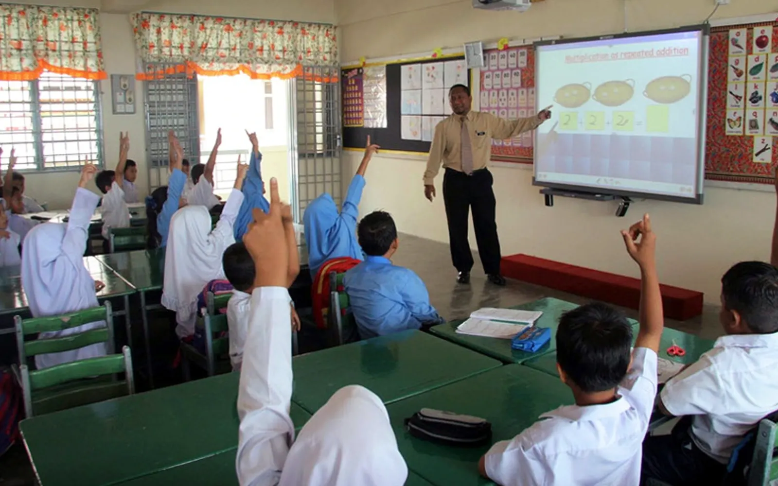 A teacher stands beside an interactive whiteboard as primary school pupils raise their hands in a classroom