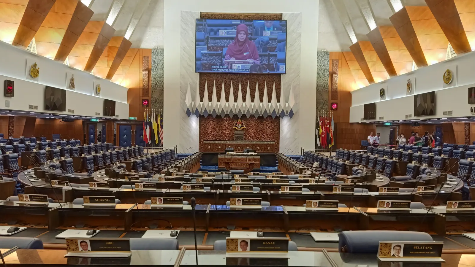 The Dewan Rakyat with rows of blue seats and a large screen above the Speaker's chair